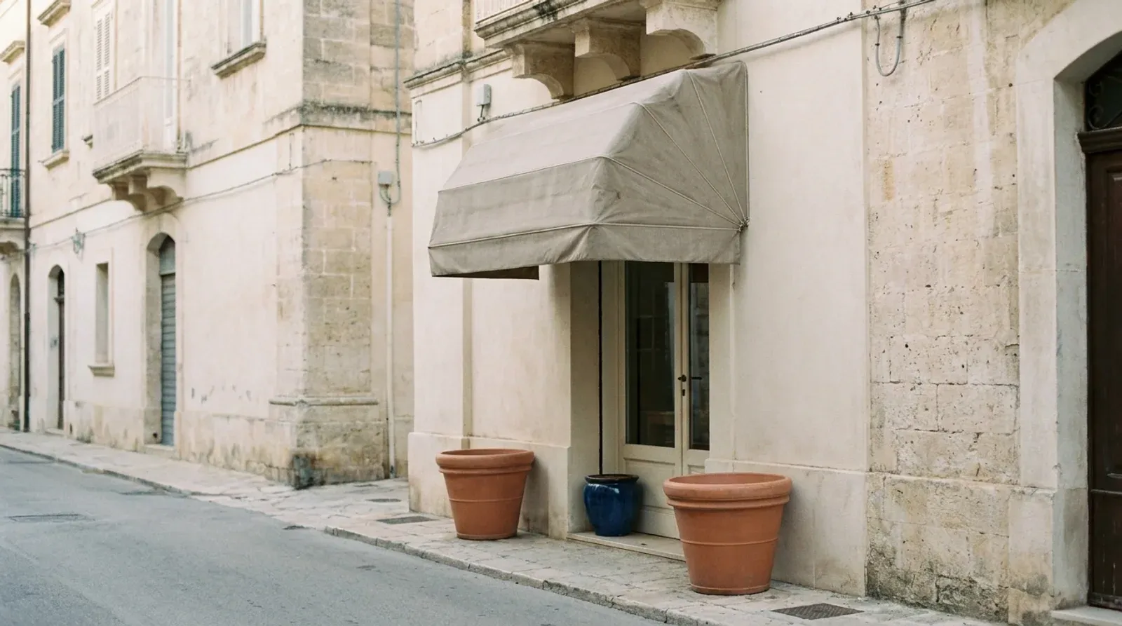 Rue méditerranéenne aux façades de pierre claire, entrée d un commerce sous un store beige encadrée de pots en terre cuite.