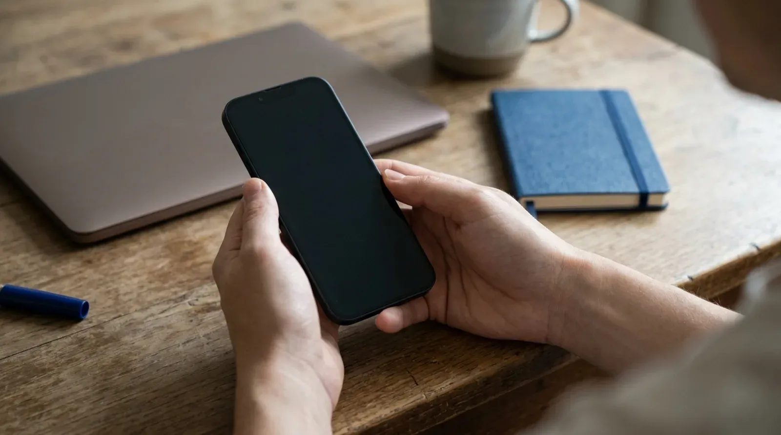 Deux mains tiennent un téléphone à l’écran éteint au-dessus d’un bureau en bois, près d’un ordinateur portable fermé et d’un carnet bleu.