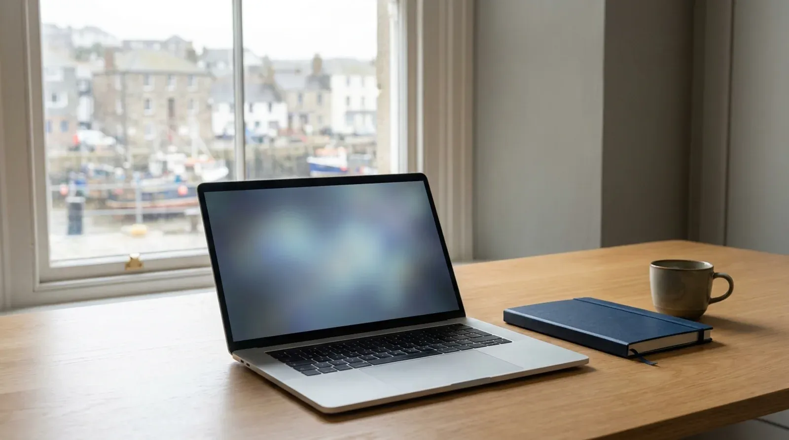 Ordinateur portable ouvert sur un bureau en bois face à une fenêtre donnant sur un port de pêche, écran vide, carnet bleu et tasse posés à côté.
