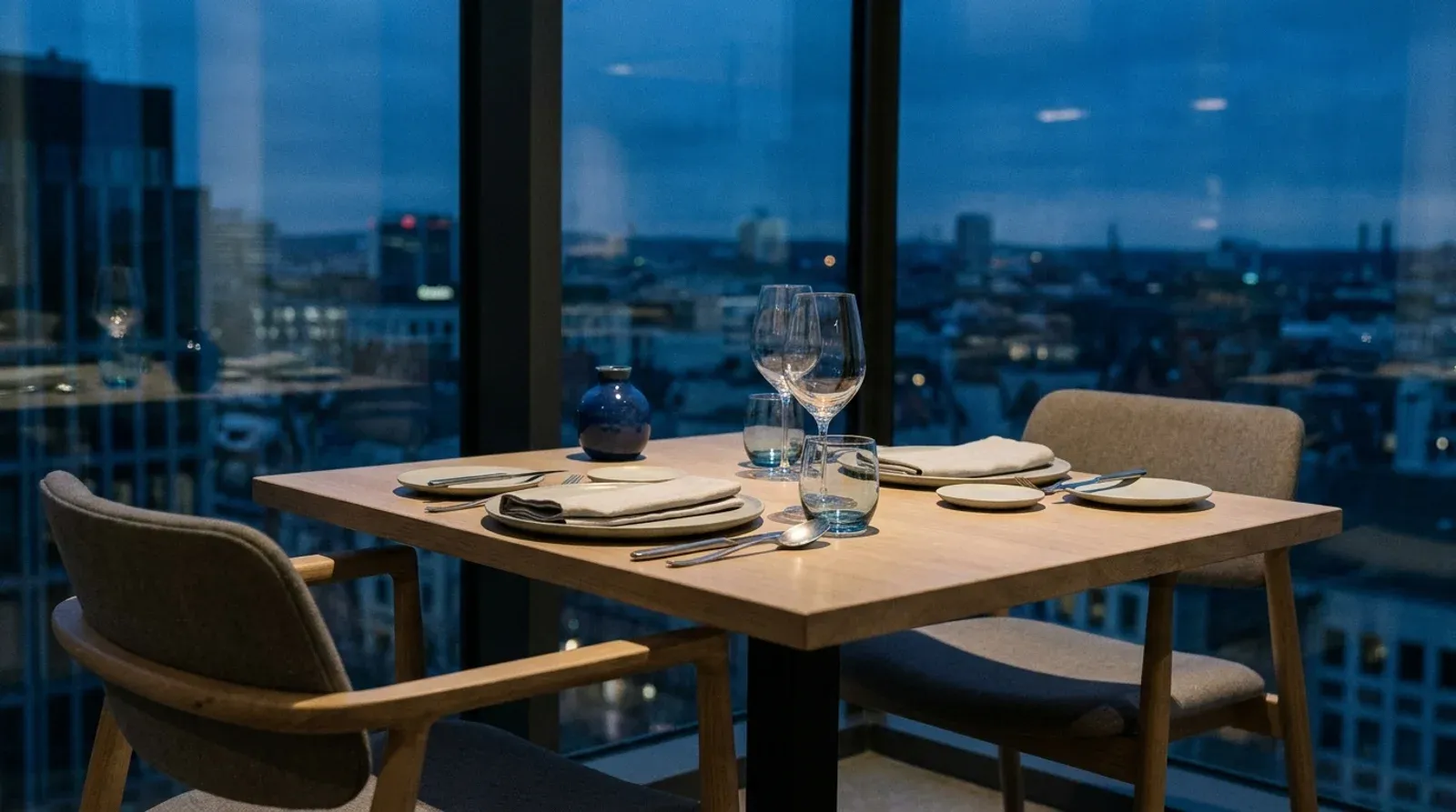 Table dressée pour deux devant une baie vitrée avec vue sur la ville au crépuscule, dans un restaurant moderne.